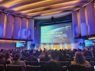 Photo : Salle de conférence moderne. Au centre de l'image, un grand écran projette des informations. La directrice générale à l'éducation, jeunesse, sport et culture de la Commission Européenne, Pia Ahrenkilde Hansen s'adresse à une assemblée derrière un pupitre.