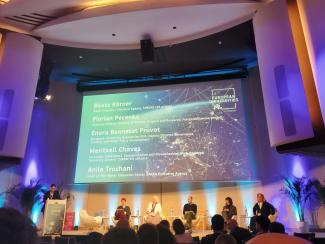 Photo : Salle de conférence moderne. Au centre de l'image, un grand écran projette des informations. 1 personne se tient debout devant un pupitre et 5 personnes sont assis devant une assemblée et semblent engagées dans une présentation ou une discussion.