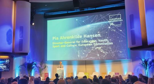 Photo : Salle de conférence moderne. Au centre de l'image, un grand écran projette des informations. La directrice générale à l'éducation, jeunesse, sport et culture de la Commission Européenne, Pia Ahrenkilde Hansen s'adresse à une assemblée derrière un pupitre.